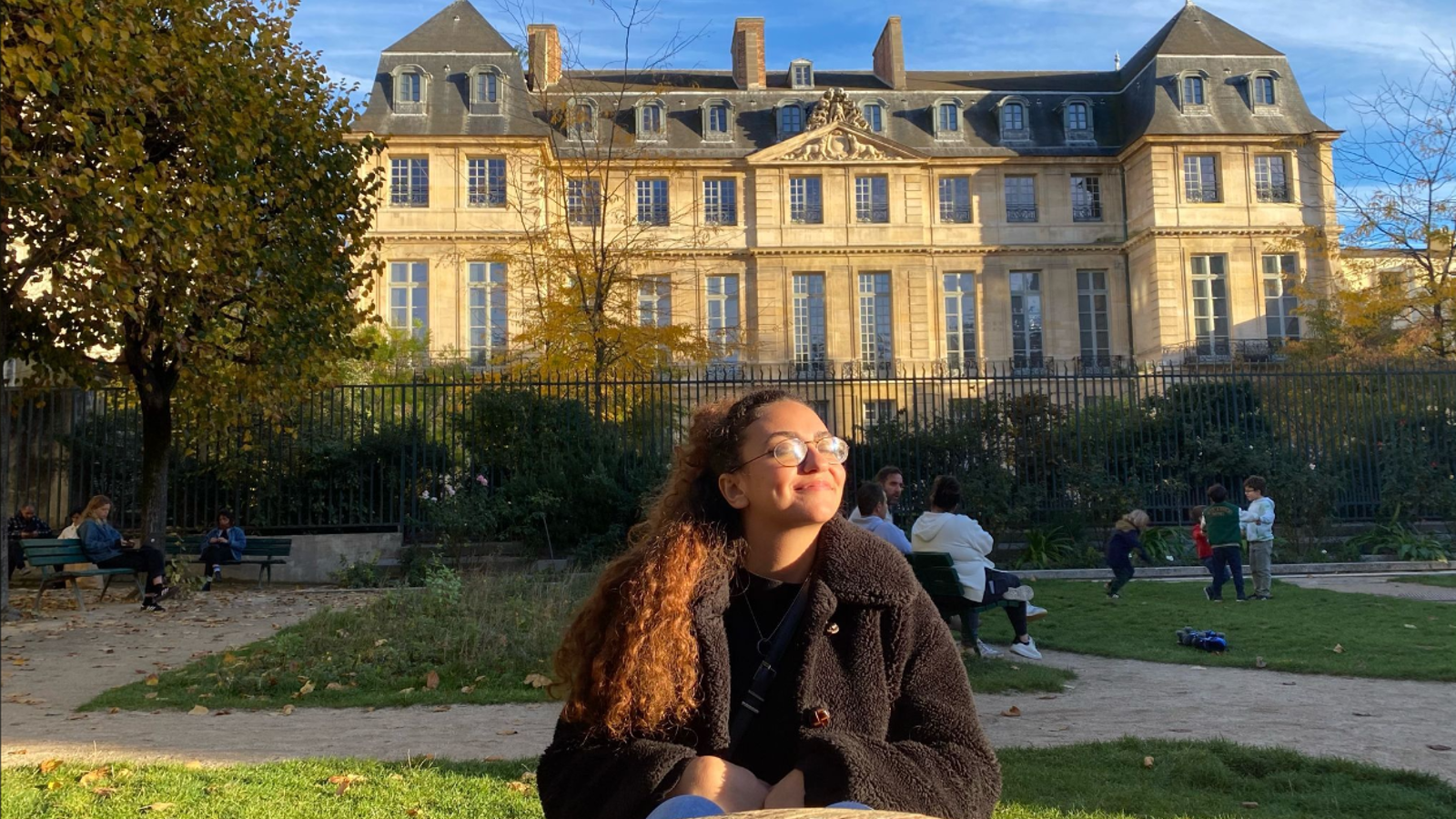a woman sitting in a park with a large building in the background with Musée Rodin in the background